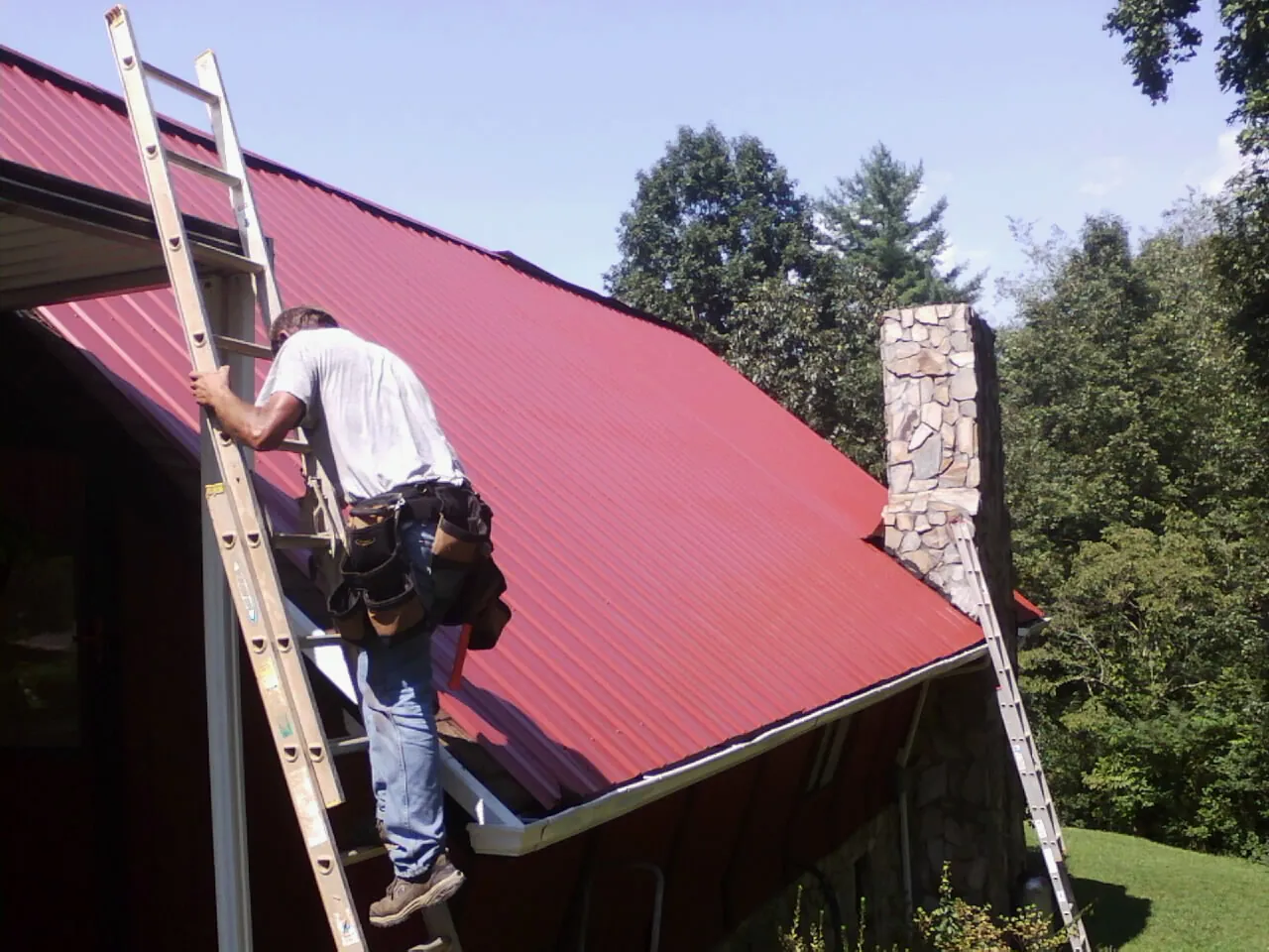 Roofer on a ladder beside a red metal roof and stone chimney — Soffit & Fascia Repair by Roofing Brothers in Lithonia